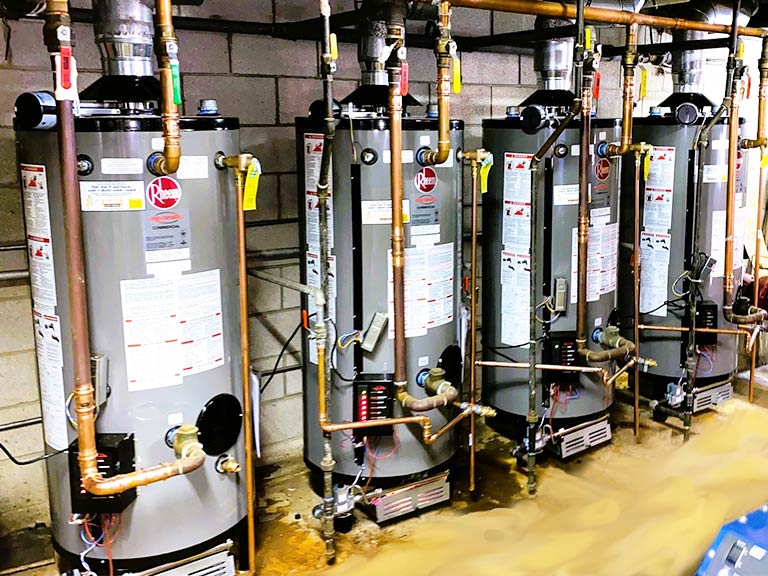 Four standard tank water heaters are lined up against a cinder block wall.