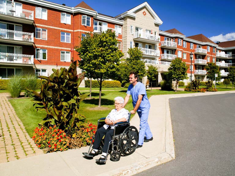 A man wearing light blue scrubs is pushing an elderly woman in a wheelchair along the sidewalk in front of a large senior living complex.