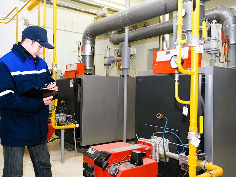 A man in a blue coat and hat inspects a boiler in a large utility space. He is holding a clipboard and pencil.