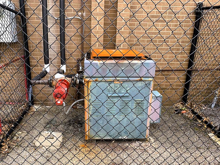 An outdoor heating boiler is shown on the ground behind a chain link fence.