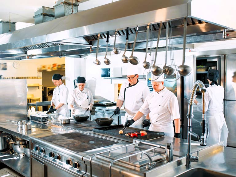 A large restaurant kitchen is shown with stainless steel appliances and counters. Several chefs wearing white jackets and hats are cooking and preparing food.