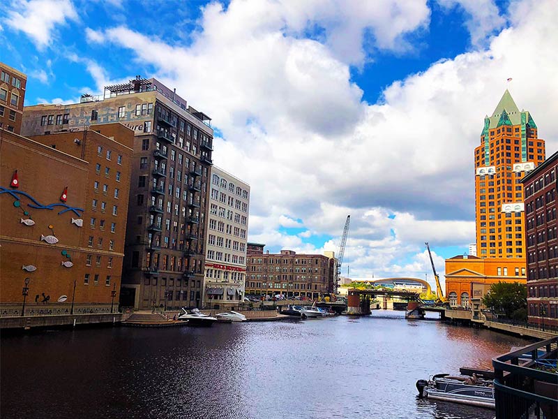Buildings line the Milwaukee River in downtown Milwaukee, Wisconsin on a sunny day with cloud-filled blue skies.