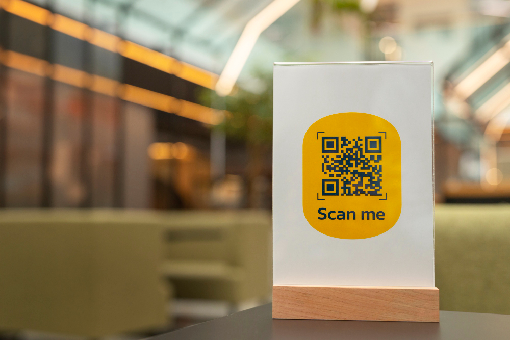 A sign with a QR code sits on a table in the foreground of a restaurant. The sign says. "Scan me."