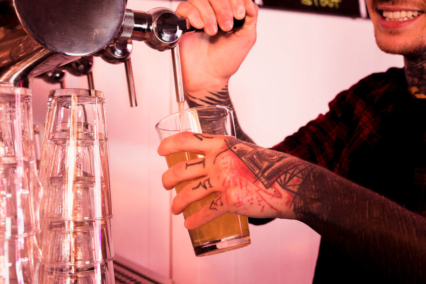 A bartender with tattooed hands pours a beer into a pint glass from a silver tap.