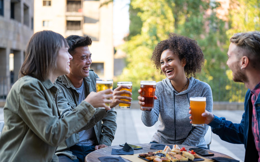 Four friends sit outside, enjoying pints of beer around a table with a plate of appetizers in the center. The friends are smiling and cheers-ing their beers.