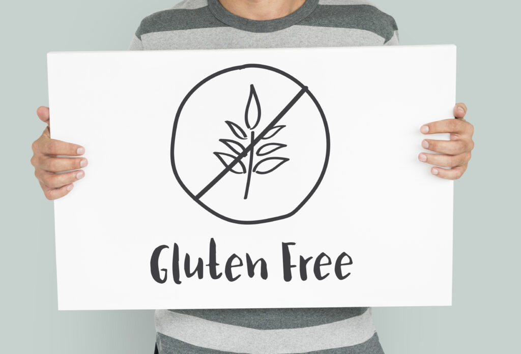 A large white sign held by a person behind it has a drawing of hops inside a circle with a line through it and the words, "Gluten Free" written underneath it. 