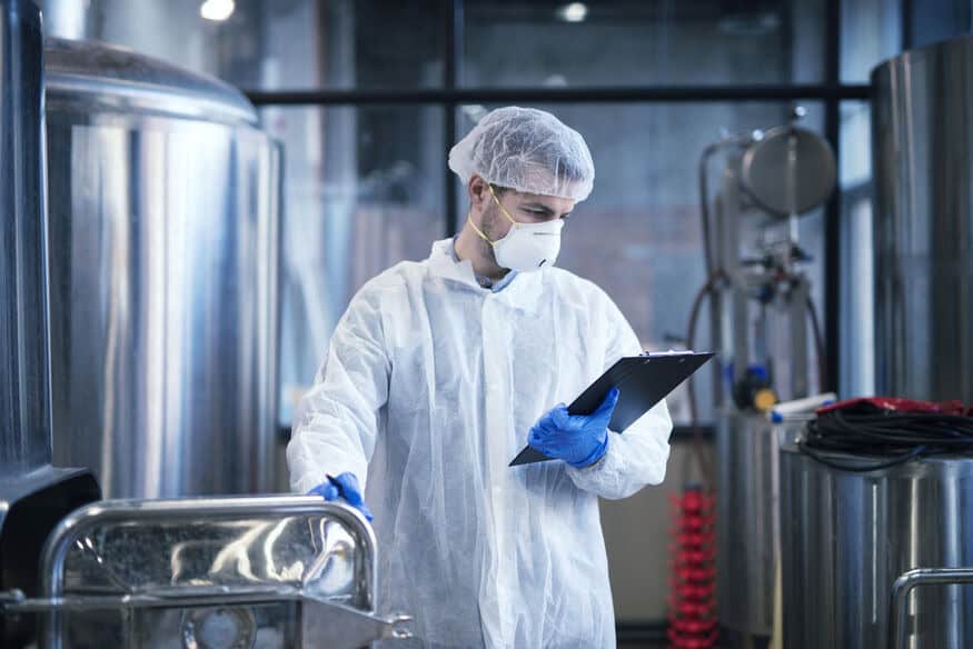 A man in personal protective equipment, wearing a mask, gloves, and a hair net, looks at the clipboard in his hand to make sure the commercial hot water system is operating properly. 